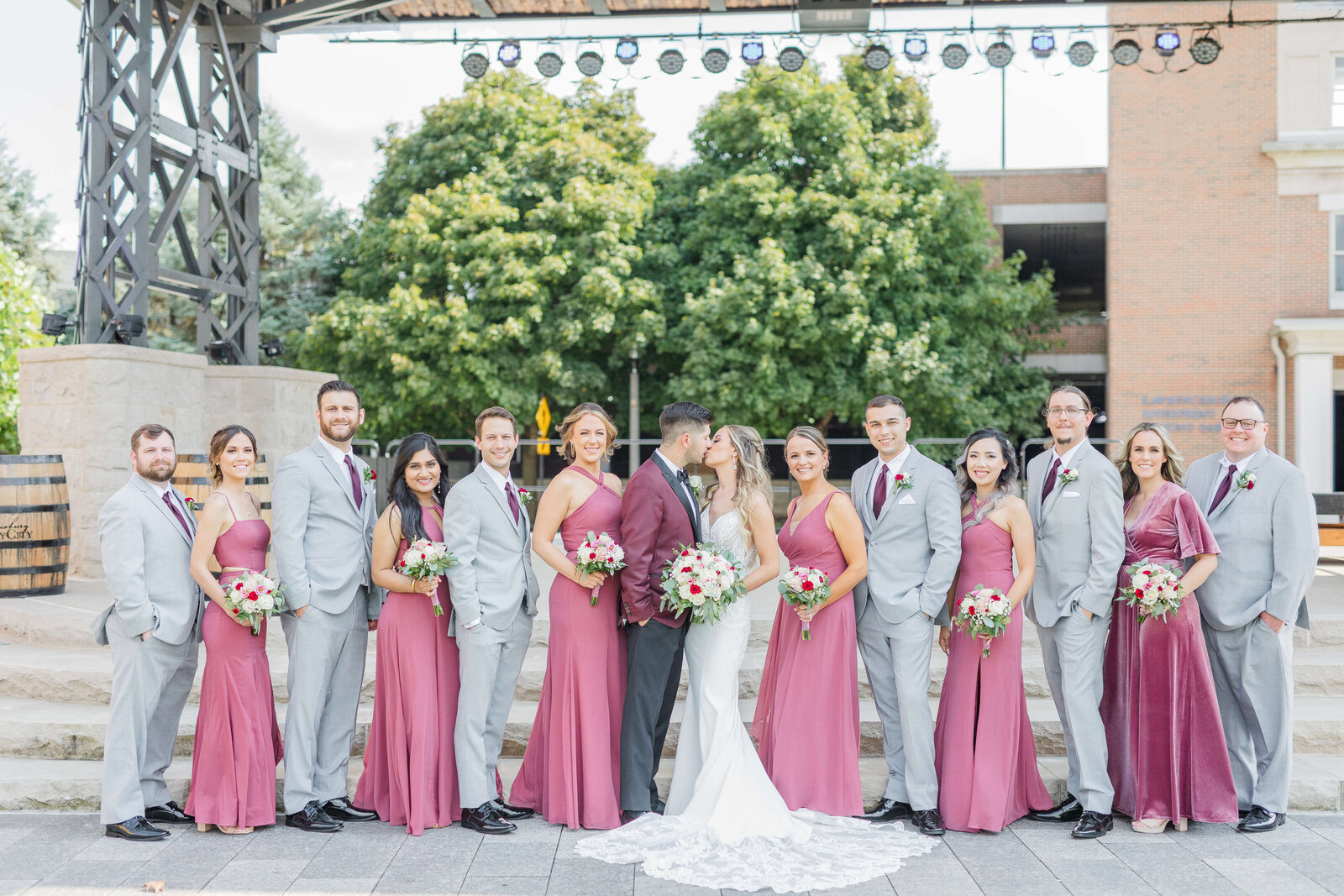Bride and groom  stand with their bridal party. There is a bridesmaid, then groomsmen, and they alternate accordingly. The bridesmaids are in rose and the groomsmen in grey.
