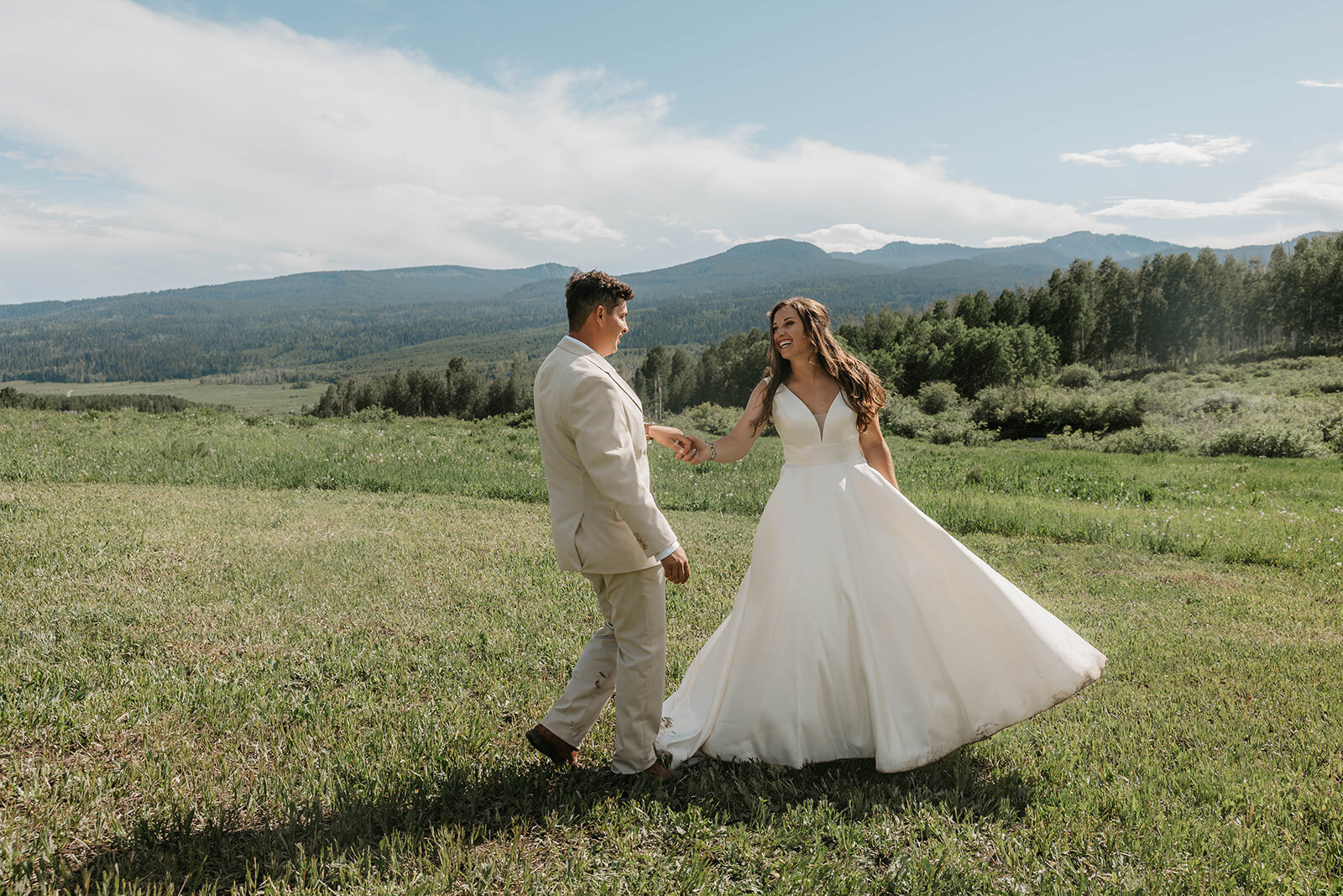 A bride and groom smile and hold hands outdoors in a grassy field, as mountains rise behind them—an intimate wedding in the Colorado mountains captured by a Colorado elopement photographer capturing real connection.