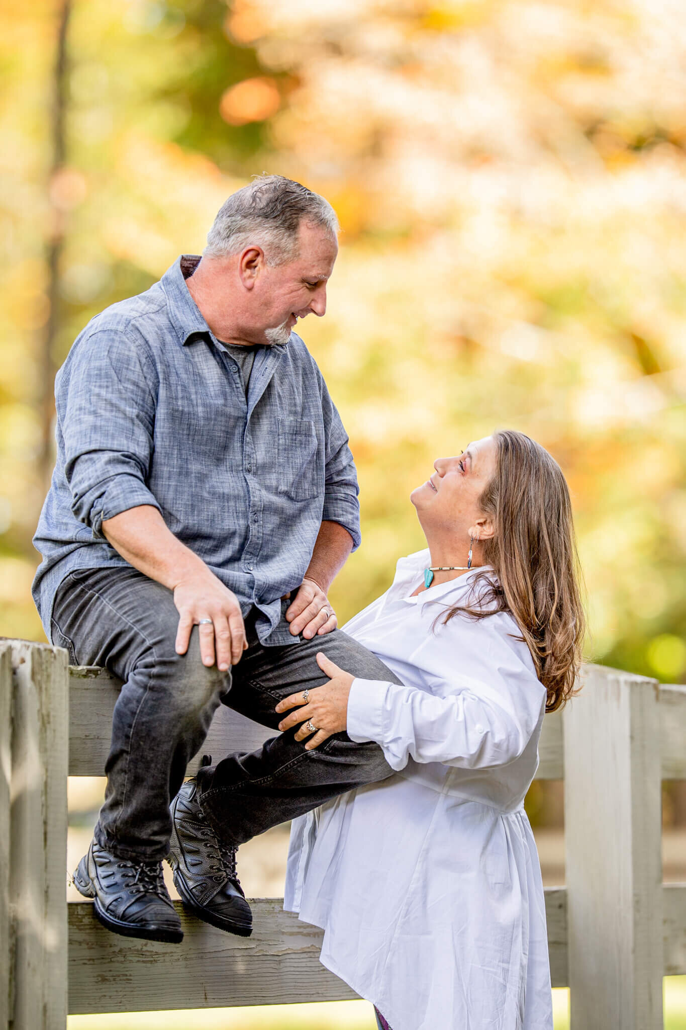 Couples photo of a male sitting on a fence weraing blue and black looking down at his wife who is standing up and looking up at him