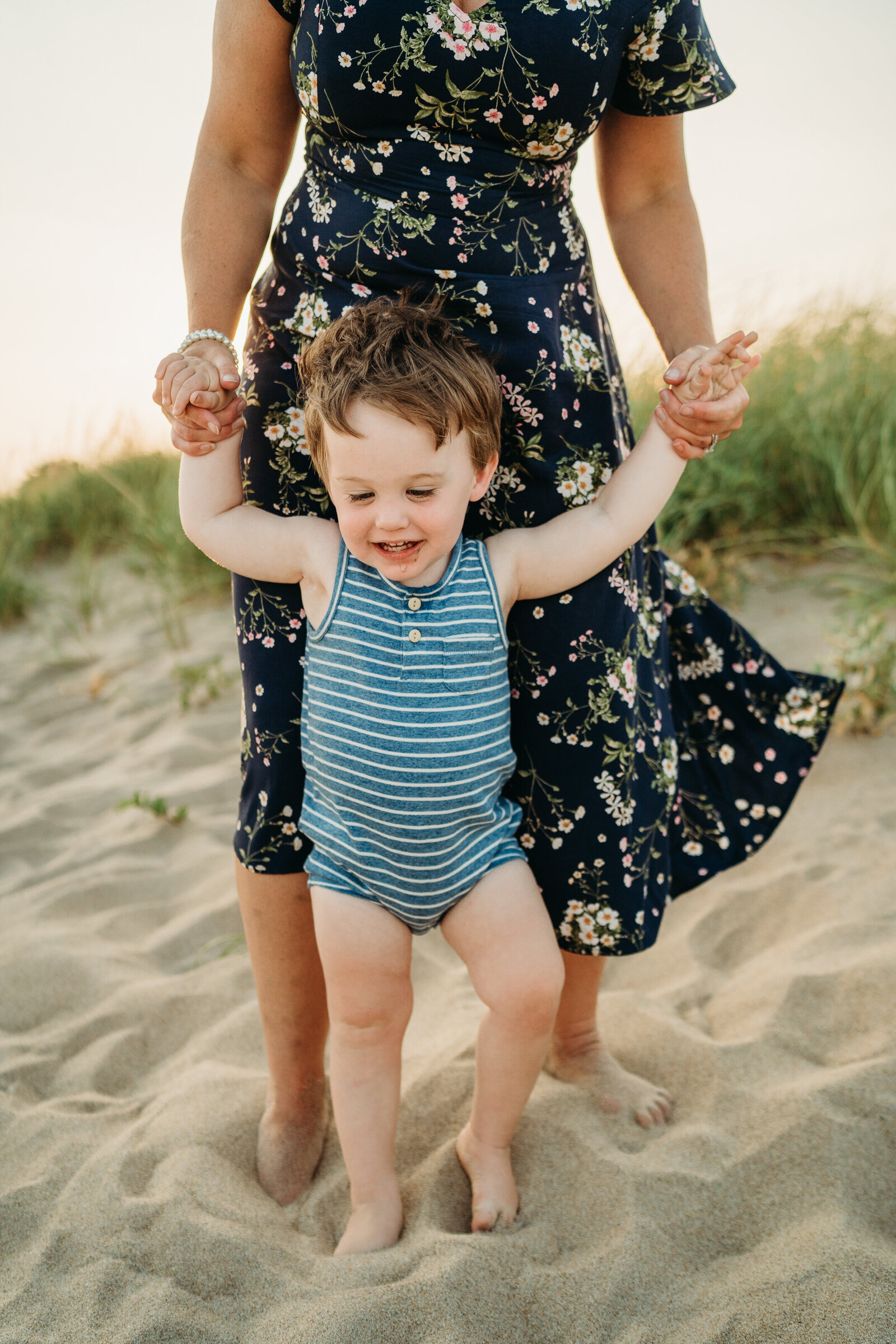 baby boy in blue striped romper walks holding onto mothers hands