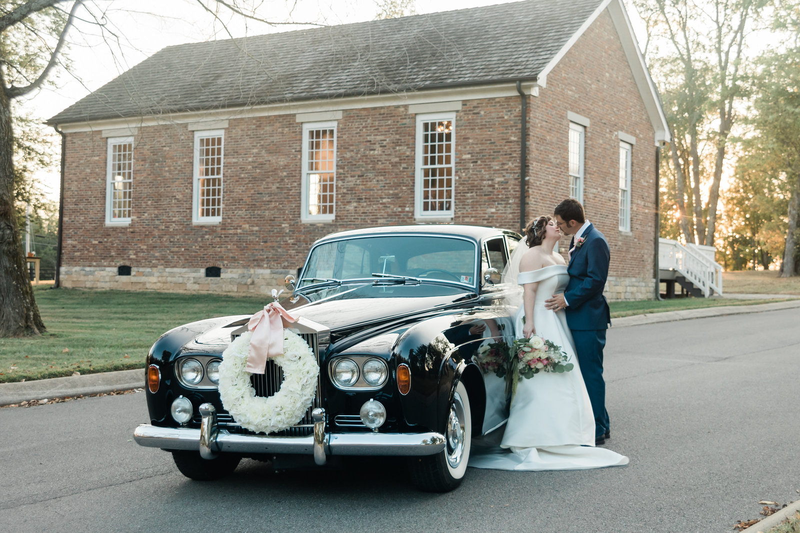 bride and groom outside of an old brick church leaning up against a vintage car as teh groom kisses teh bride in Knoxville Tennessee