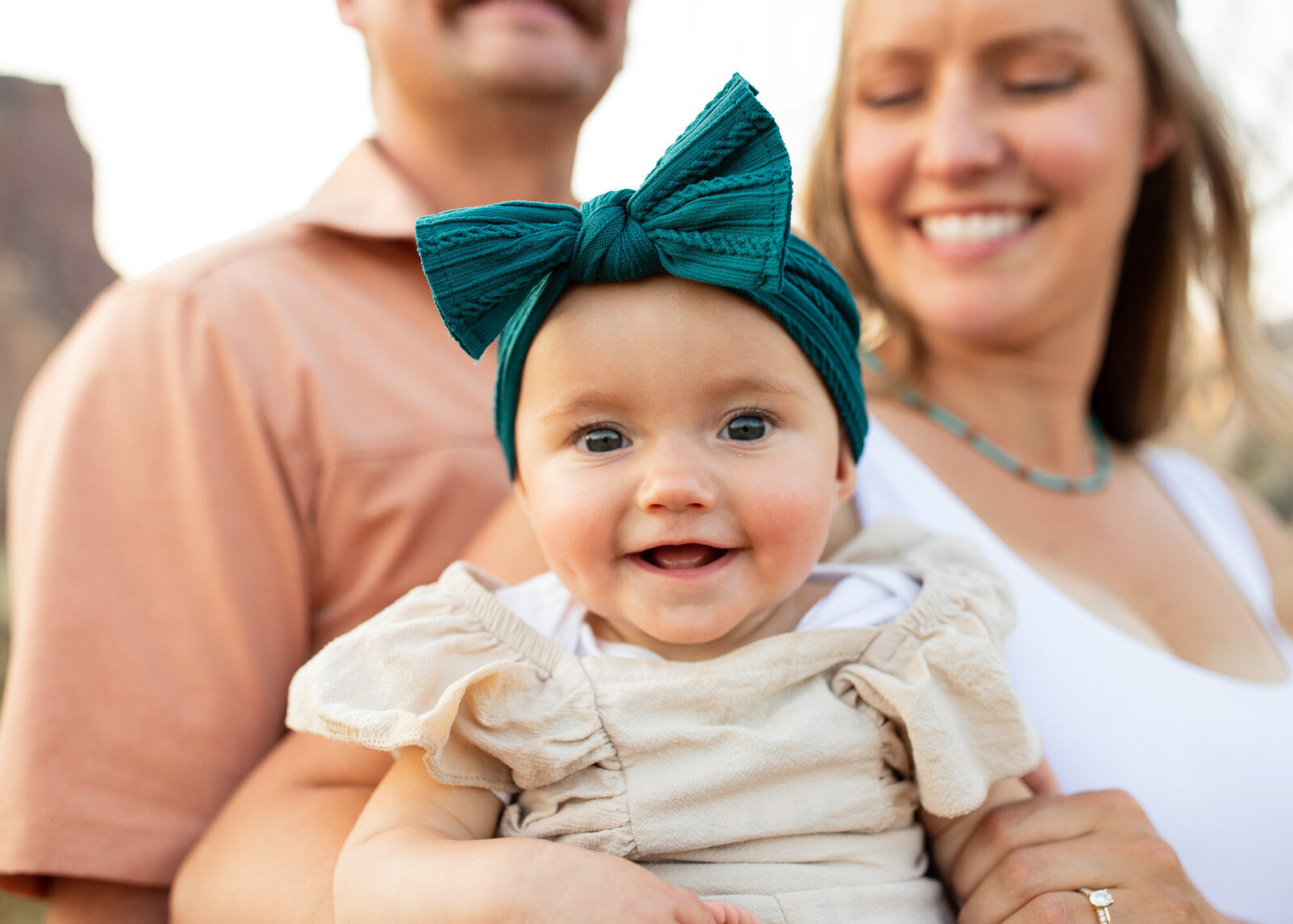 Baby smiling with big jade colored bow on while parents hold her.