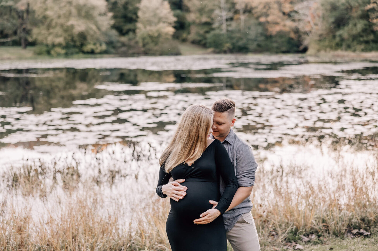Pregnant woman with blond hair in black dress poses with her husband in front of Sawmill Pond in Ledyard, CT