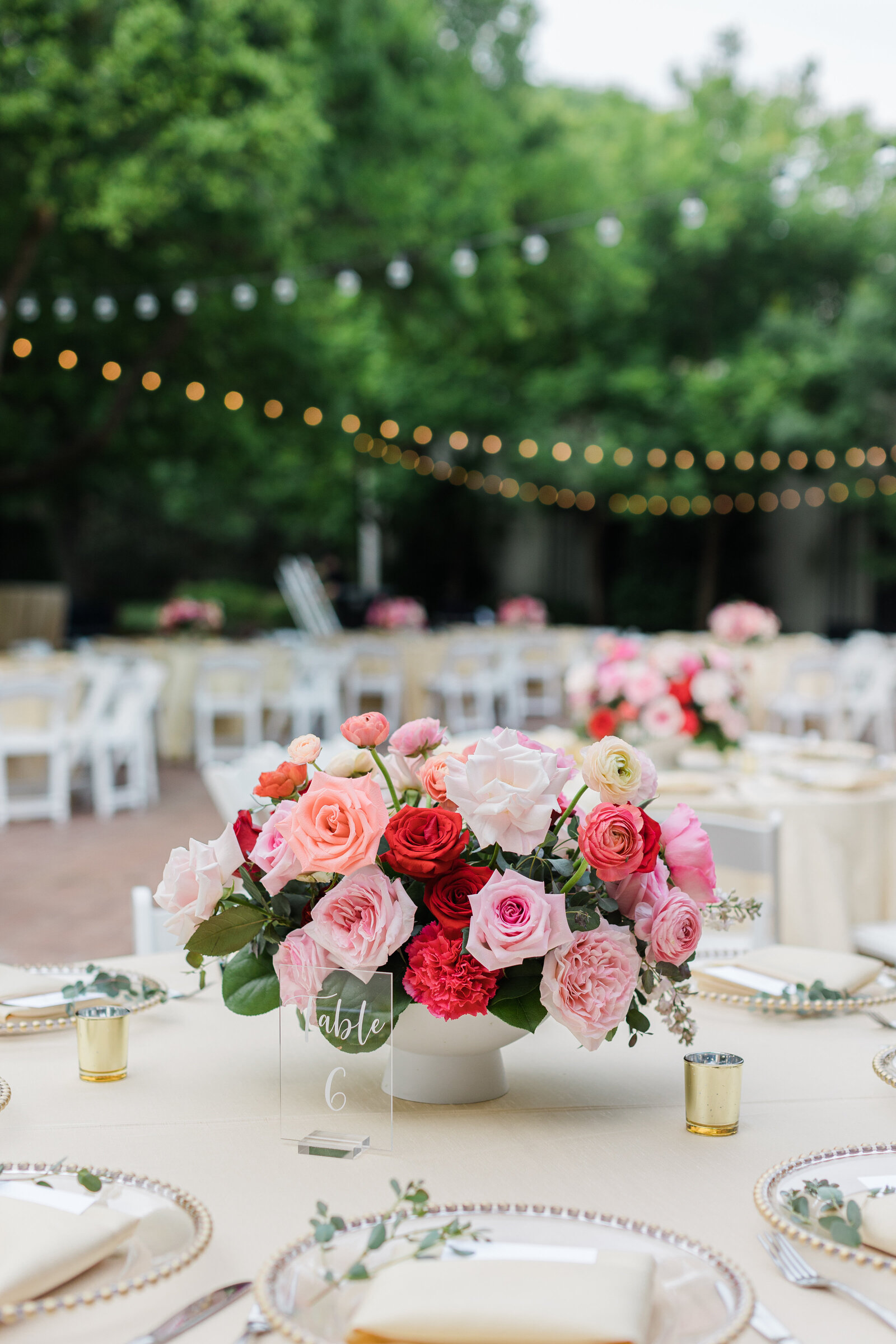 A detail shot of a wedding reception table centerpiece at the Dallas Arboretum in Dallas, Texas. The centerpiece is composed of many pink and red roses and is surrounded by a table number sign, plates, cutlery, napkins, and candles. Similar tables can be see out of focus in the background.