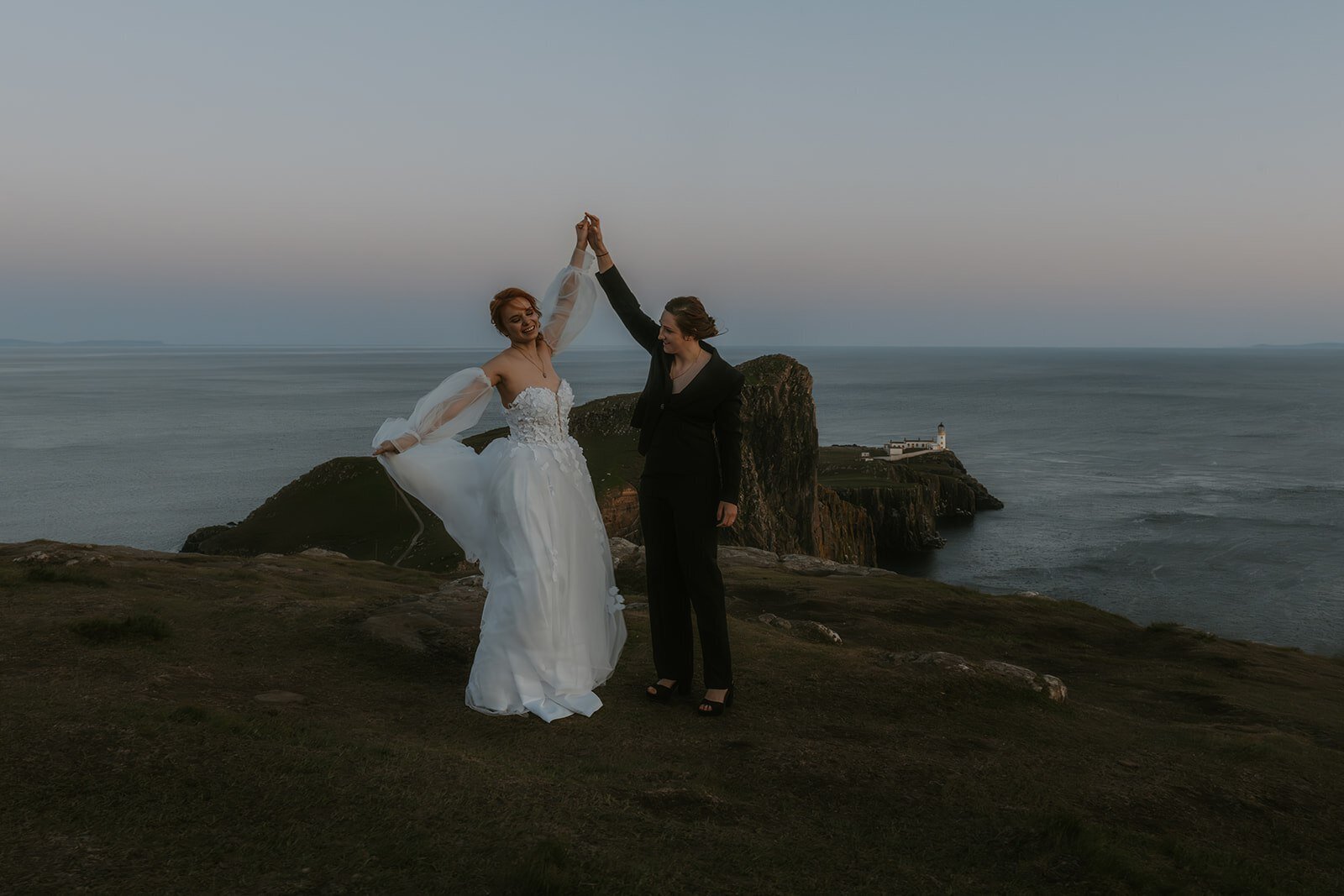 Two women, one in a wedding dress and one in a suit, dance joyfully on a cliff by the sea at sunset—capturing the spirit of a photography retreat in New Zealand for creatives and connection.