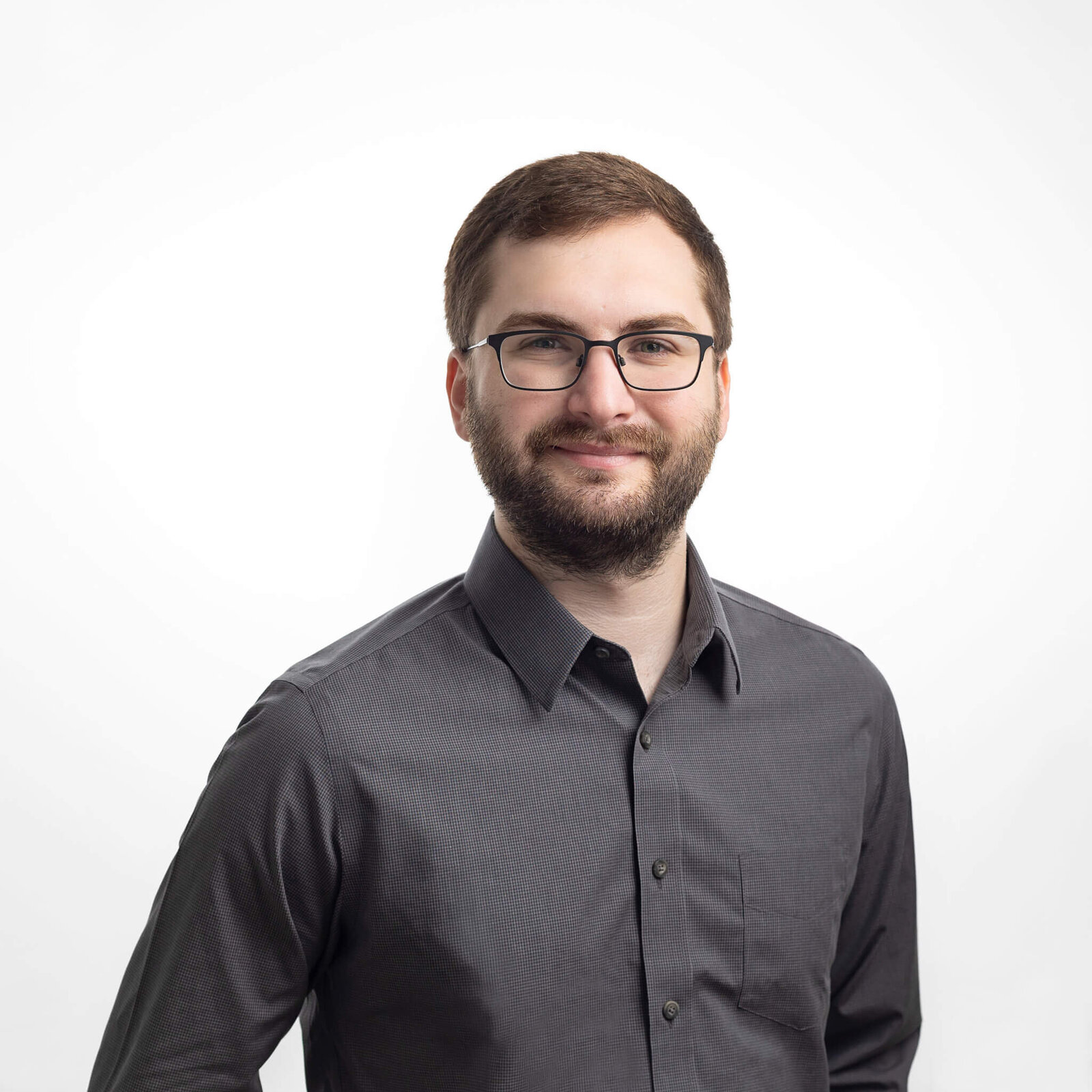 Young man is wearing a grey button shirt and glasses over a white backdrop in a studio headshot session by Ingrid Barnhart Photography in Austin Texas