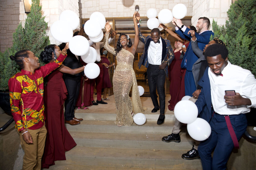 bride in a gold dress and groom holding their hands up and leaving the reception with guests tossing white balloons