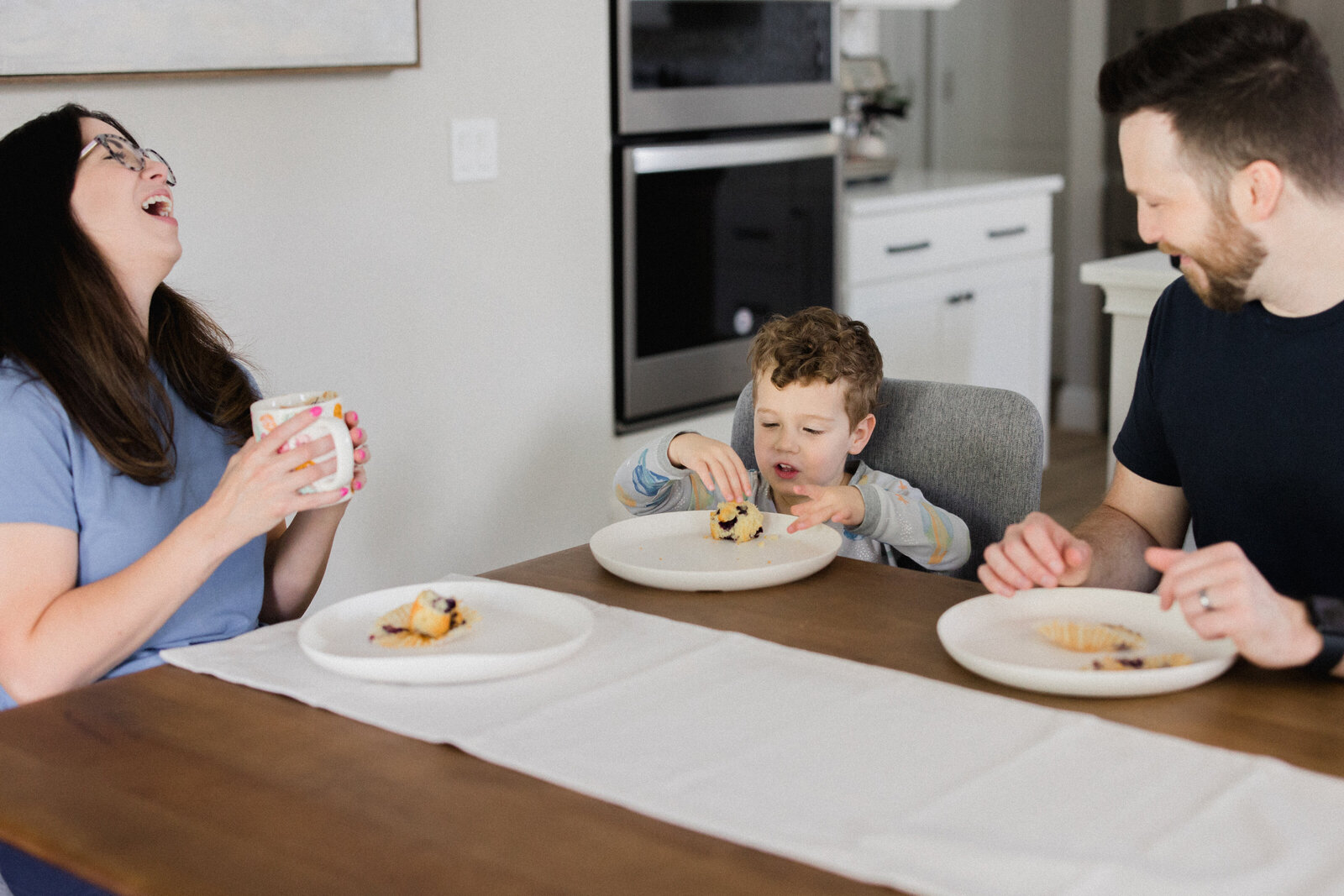 Mom laughs as she eats breakfast at the dining room table with husband and son