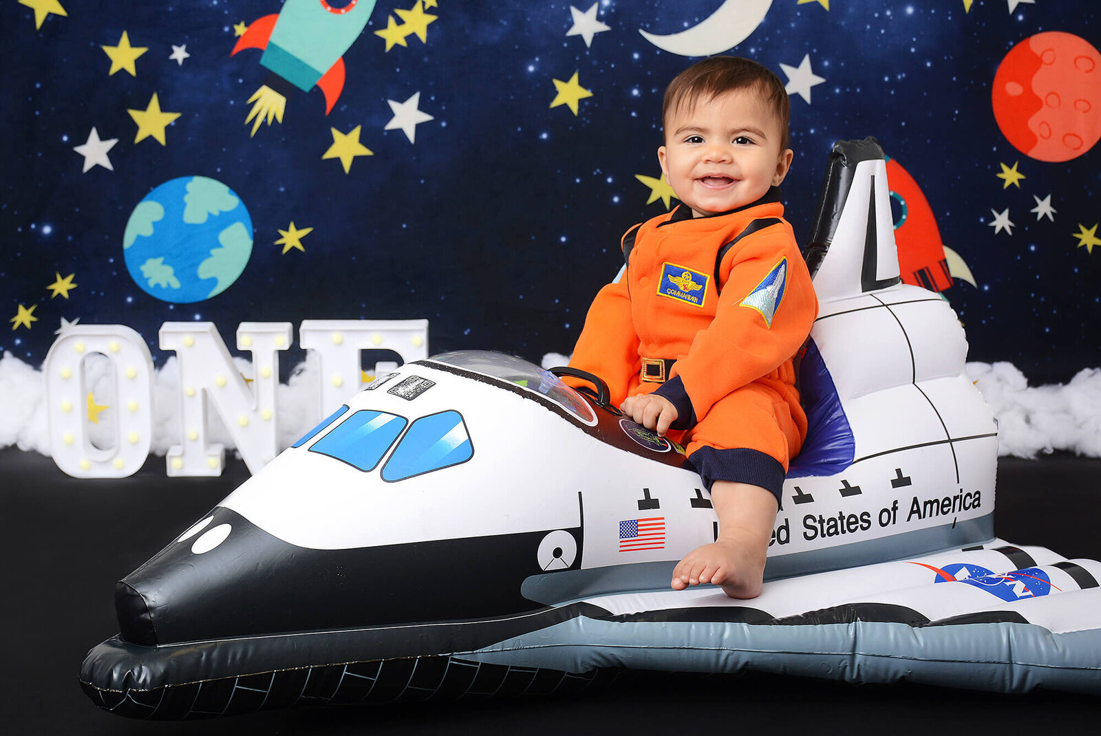 boy smiles while on a rocket ship prop with small planets behind him and the word one