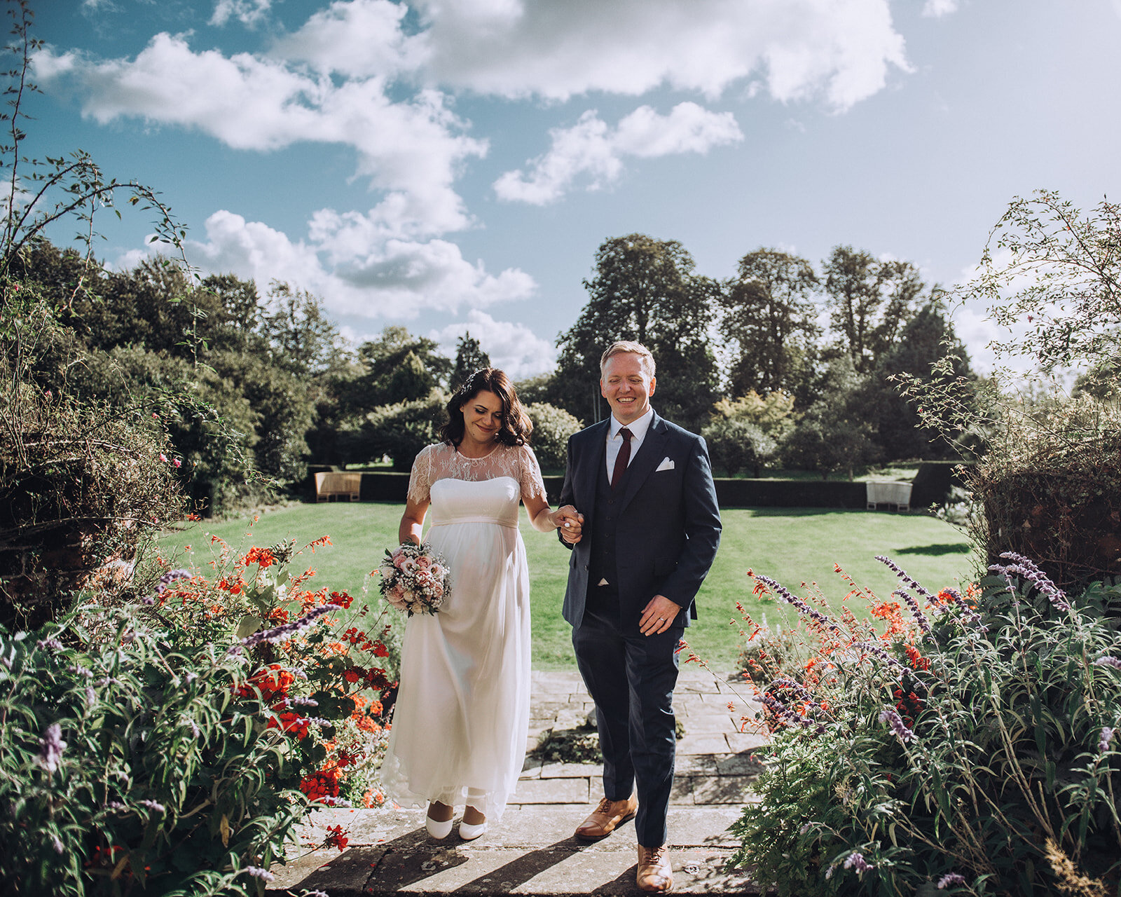 Couple hold hands and smile as they walk between flowers at their wedding at Childerley