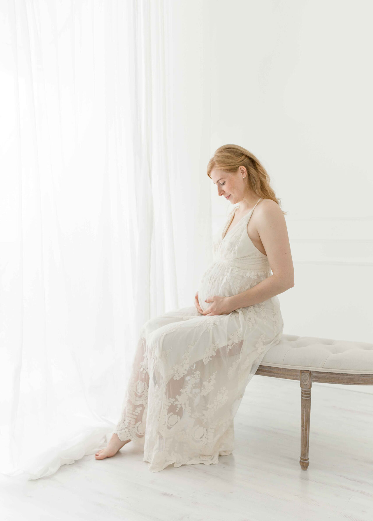 Babybauchshooting luxuriös in weiß . Schwangere Frau im langen weißen Spitzenkleid am Fenster sitzend.