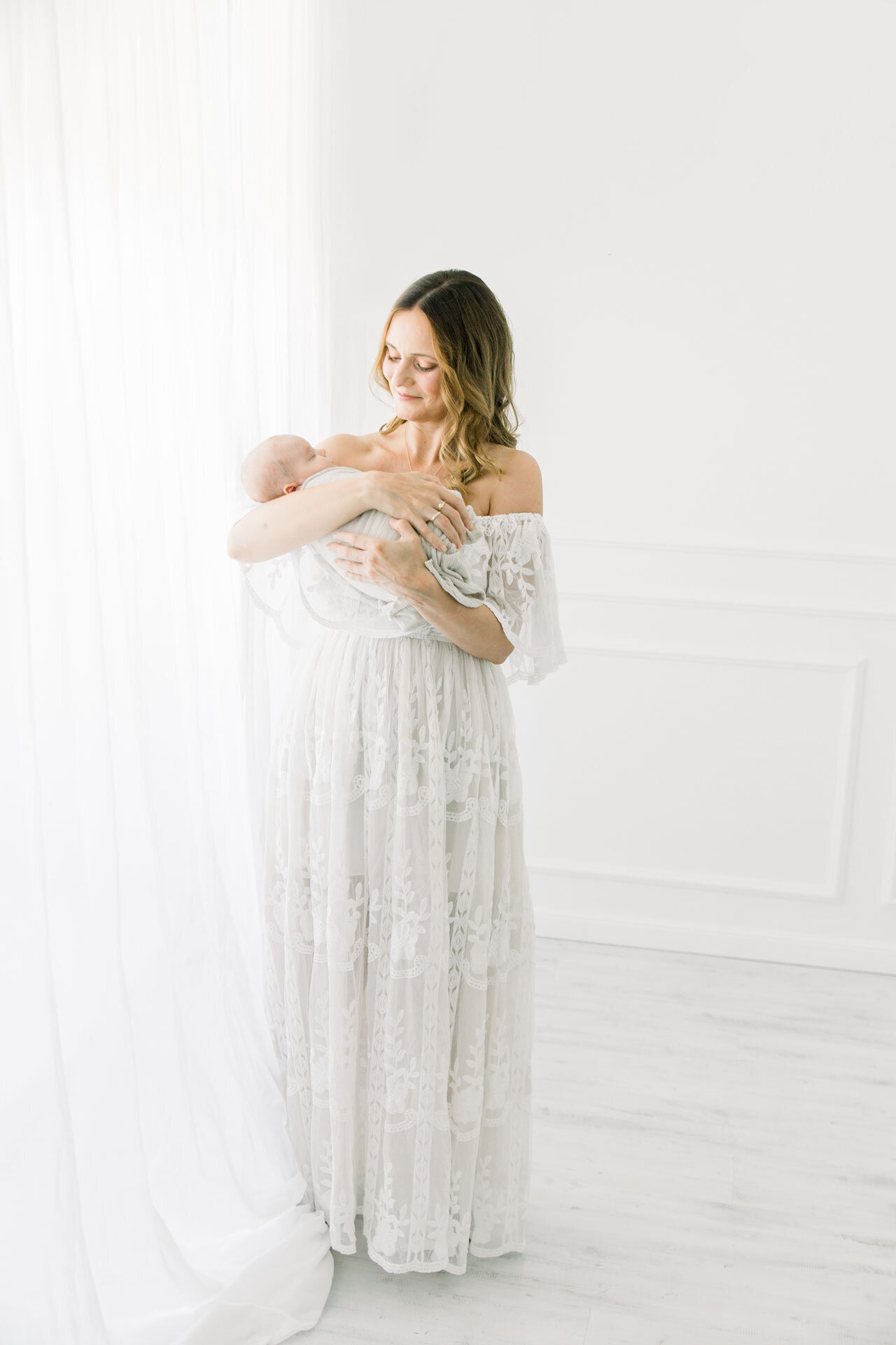 Babyshooting: Mama in langem weißen Spitzenkleid vor weißer Gardine hält ihr schlafendes Neugeborenes im Arm.