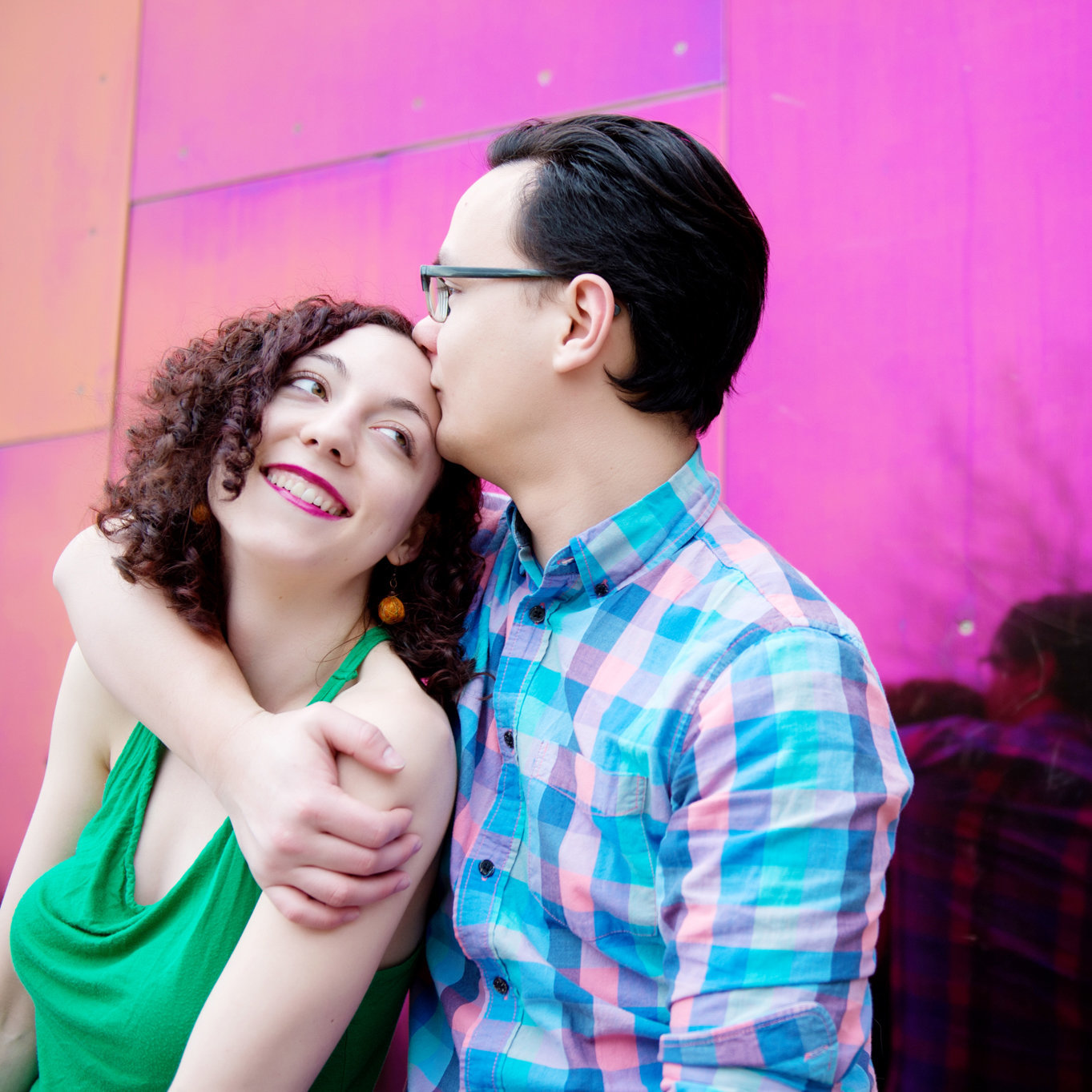 a man kisses a woman's forehead in front of a bright pink shiny wall