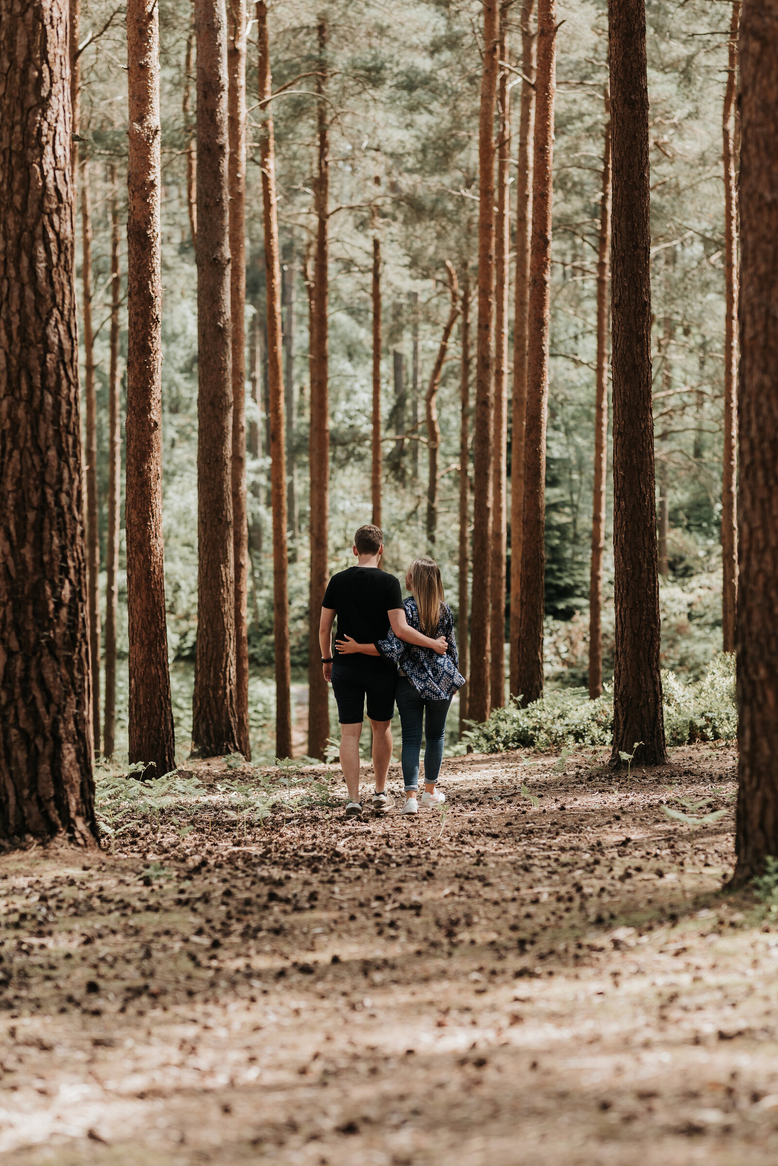 Couple walk with arms around each other along woodland pathway, through tall trees