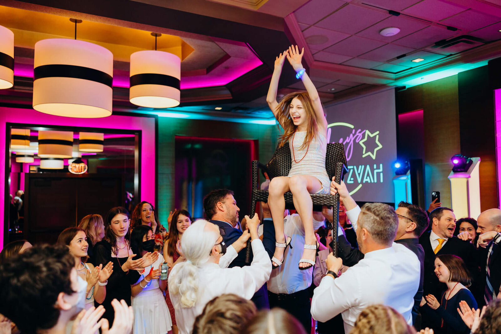A happy teen girl in a silver dress laughs while being lifted in a chair on the crowded dance floor in some Bellevue Bar and Bat Mitzvah Photography