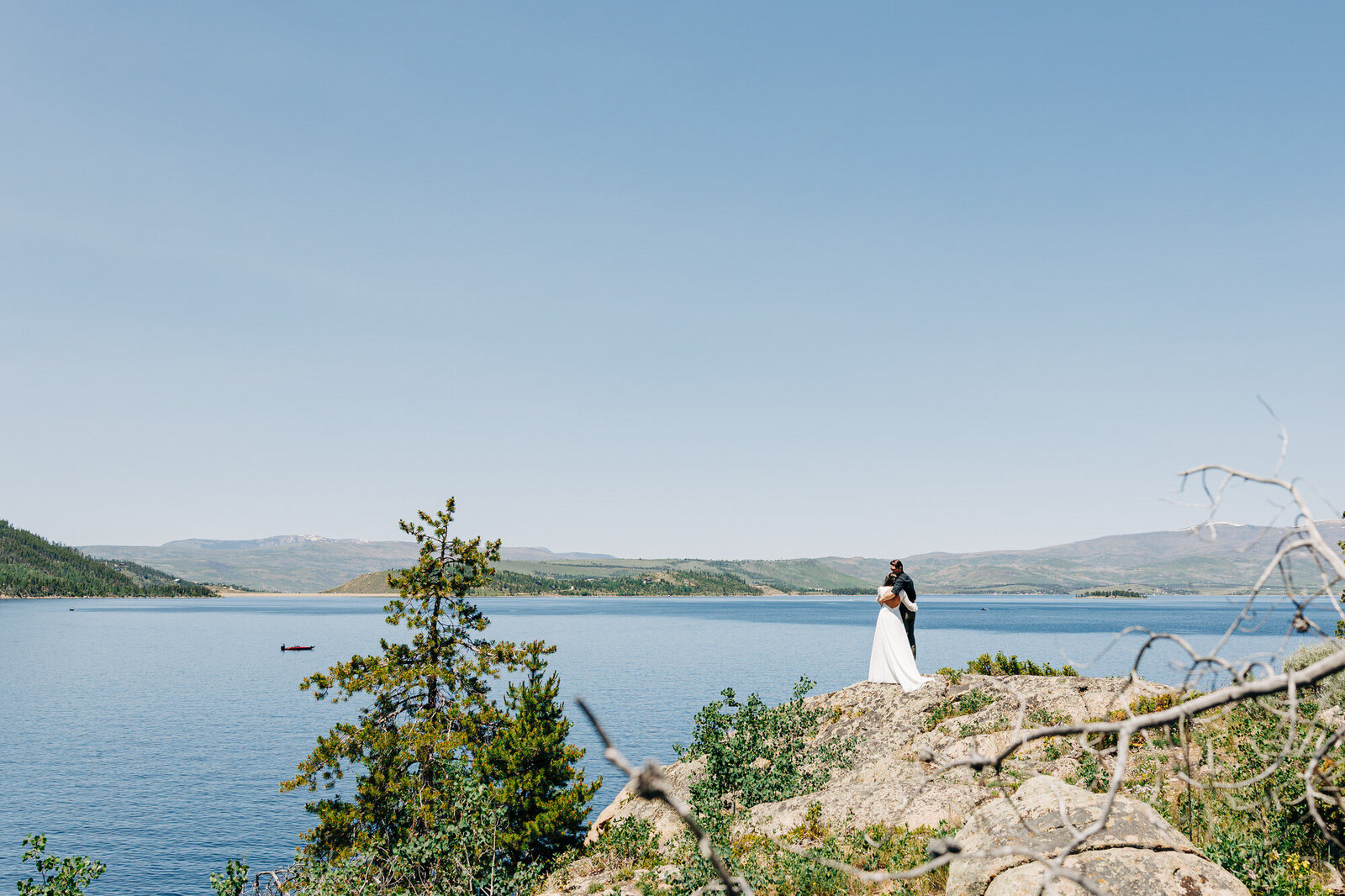 A couple in wedding attire embrace on a rocky cliff overlooking a large lake, with mountains and a clear blue sky in the background. Captured by a Colorado Wedding Photographer, a boat is visible on the water in the distance.