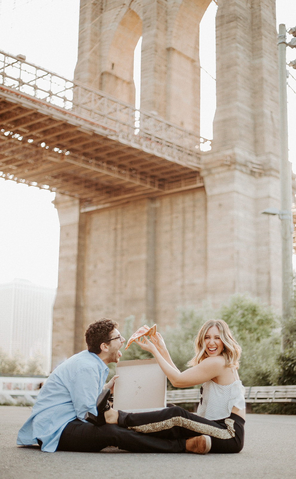 Woman feeding pizza to her boyfriend in front of a bridge while sitting on the ground during their engagement session