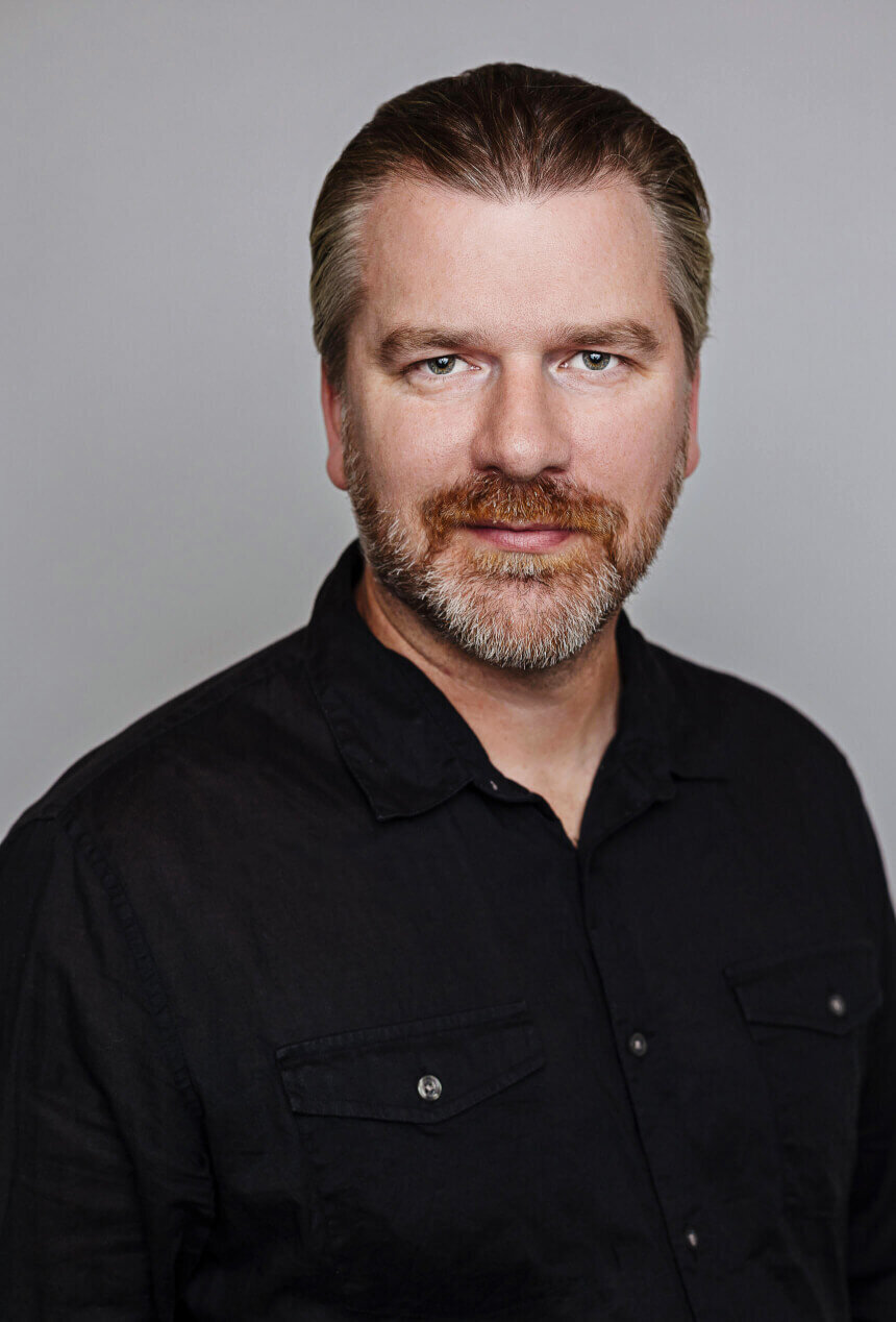 man wearing a black shirt and a beard looks very professional in a studio headshot for his company
