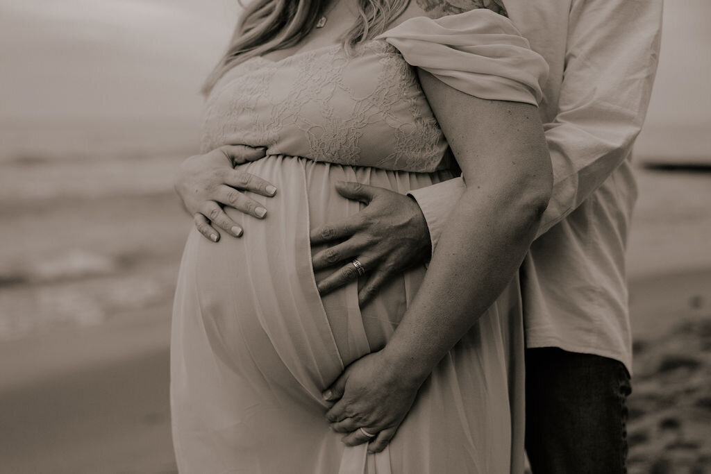 Moody black and white vintage looking maternity photoshoot by Morgan Ashley Lynn Photography in Milwaukee, WI on the beach of Lake Michigan. The man is standing behind the woman with his arms around her baby bump