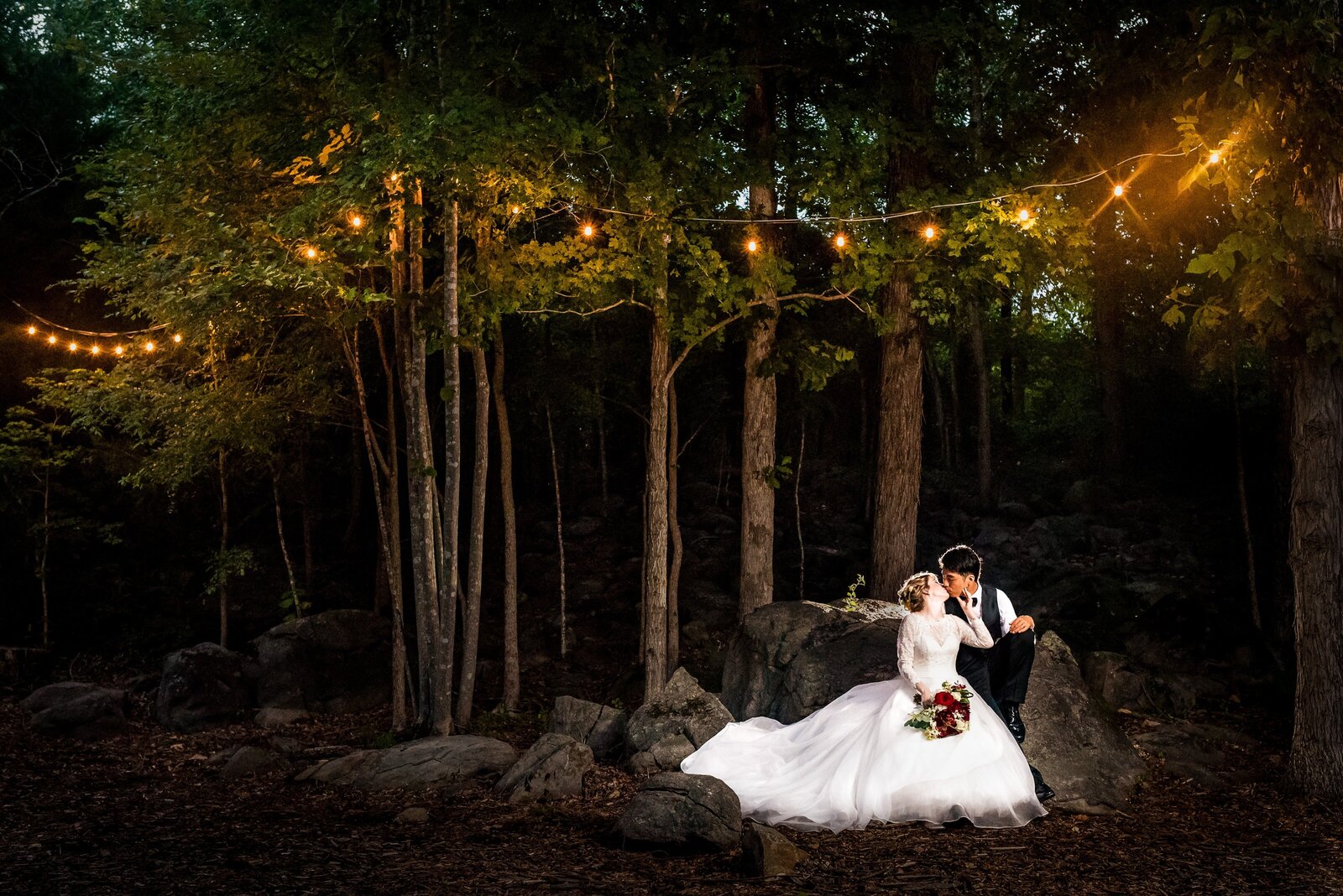 wedding couple poses on rocks with trees in the background that are strung with twinkle lights