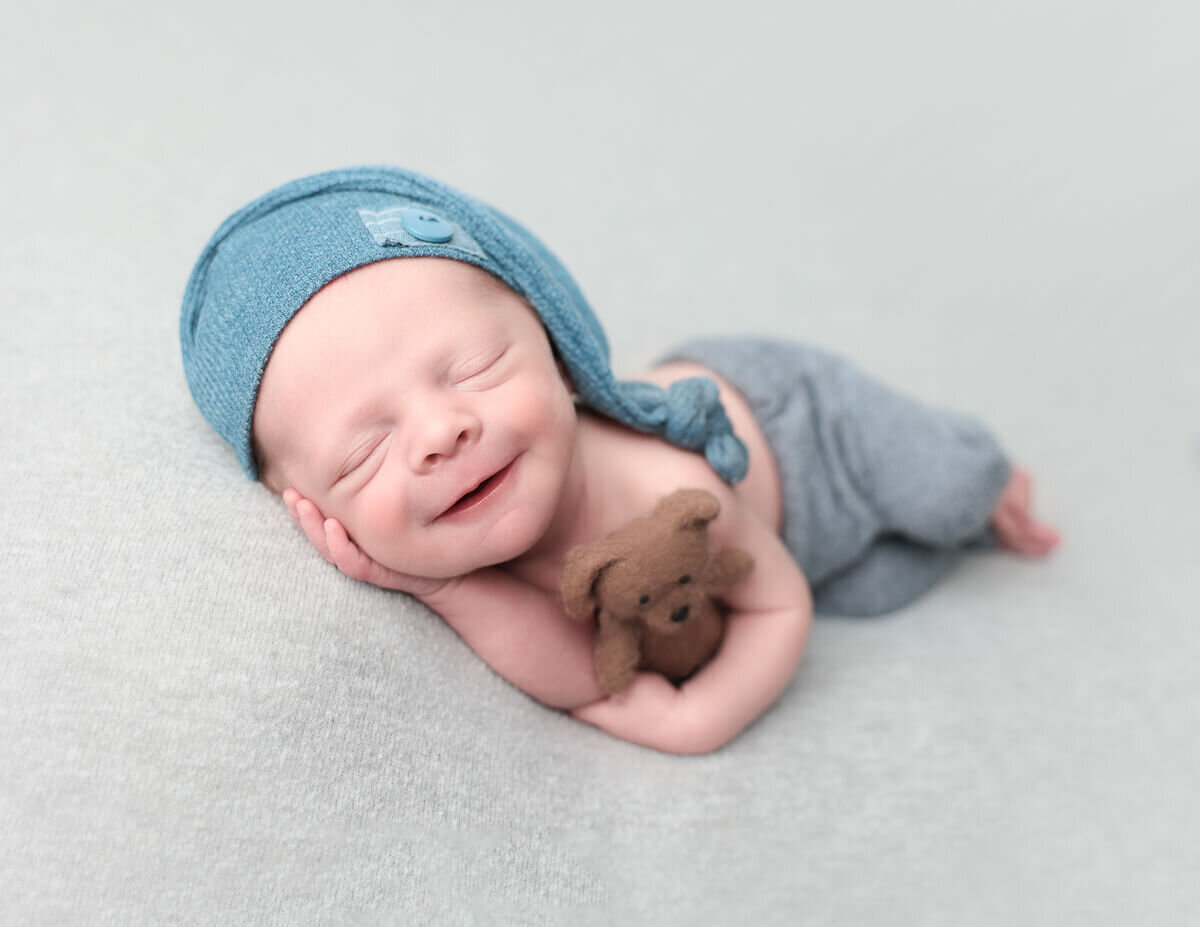 A newborn baby boy smiles while holding a teddy bear, wearing a cute sleepy cap.