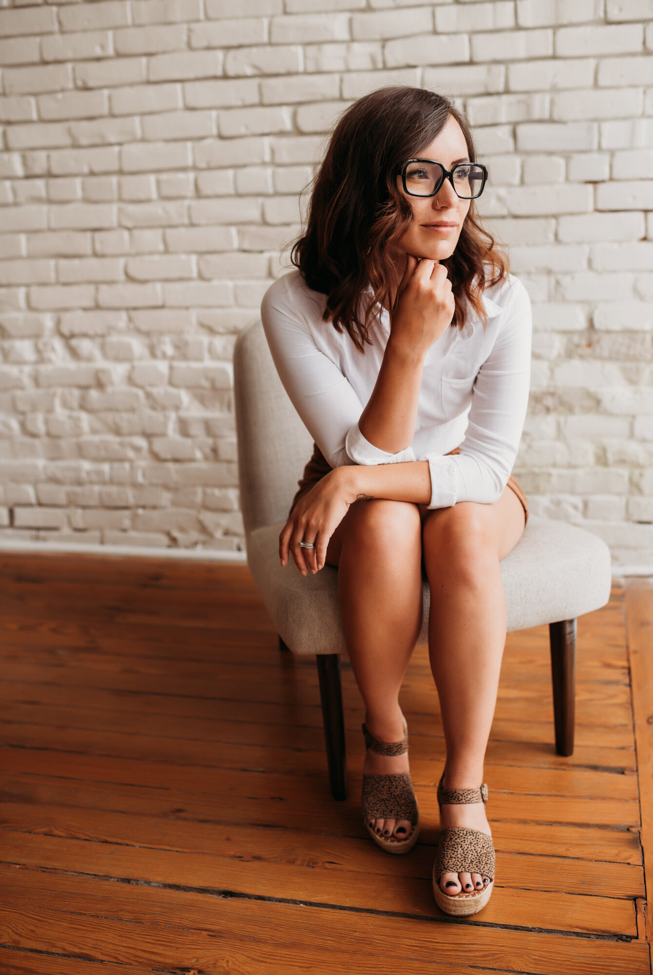 Branding Photographer, a woman with glasses sits in a chair near a brick wall inside