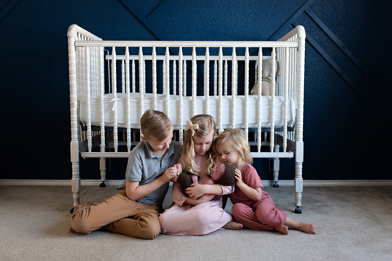 Three siblings cuddling their newborn brother while sitting on the floor of his nursery room.