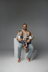 Father sitting on a white studio cube holding his two young children against a soft gray backdrop, timeless studio family portrait captured by Kaitlyn Dawn Photography, professional studio family photographer.
