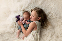 Sweet portrait of a big sister cuddling her newborn sibling while lying on a soft white rug, taken at Kara Reese Photography’s Waukesha studio serving Oconomowoc and New Berlin.