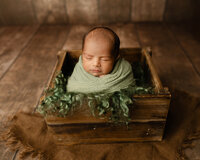 Newborn baby boy wrapped in a soft green swaddle and posed in a rustic wooden crate during a cozy studio newborn photography session by Kaitlyn Dawn Photography.