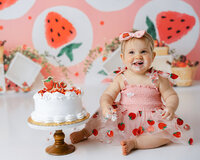 Baby in strawberry-themed dress enjoying cake smash with watermelon backdrop. Photograph by Yucaipa cake smash photographer Kaitlyn Dawn Photography.