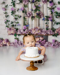 Baby leaning forward to taste a white cake during a cake smash session with a purple floral background. Photograph by Yucaipa cake smash photographer Kaitlyn Dawn Photography.
