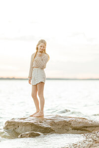 Beautiful soft sunset photo of a girl standing on a rock on the side of a lake as she looks over her shoulder to pose for the photographer.