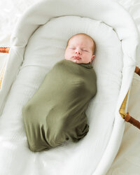 Sleeping newborn baby swaddled in olive wrap resting in a bassinet during an in-home lifestyle newborn photography session.