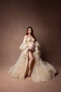 Studio maternity portrait of a pregnant woman in a flowing champagne tulle gown gently holding her baby bump, photographed by Yucaipa maternity photographer Kaitlyn Dawn Photography.