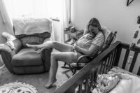In Monticello, Illinois, a new mom sits quietly in the nursery with her newborn tucked under her shirt for skin-to-skin bonding during a lifestyle newborn session. - A portrait experience by Nature's Child Photography.