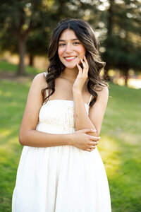 Smiling high school senior girl posing in a park wearing a white dress, Inland Empire outdoor senior portrait by Kaitlyn Dawn Photography.