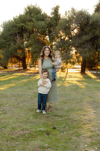 Mother standing in a sunlit park holding her baby while her young son stands beside her, warm golden hour family portrait captured by Kaitlyn Dawn Photography.