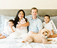 Family portrait of parents, two young brothers, newborn baby, and family dog together on a bed during an in-home newborn session.