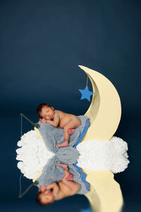 A newborn baby peacefully sleeps on a crescent moon prop, nestled among fluffy clouds. The baby is wrapped in a soft, blue blanket and lies against a dark blue backdrop, with a small blue star hanging above. Captured by a Jacksonville newborn photographer, this serene scene is truly magical.