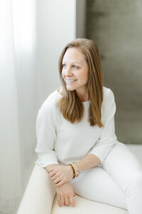 Light filled in studio professional headshot photo of a woman sitting down on a chair while she looks out of the window smiling.