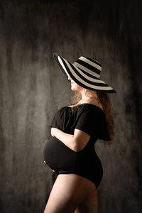 A pregnant woman stands sideways against a gray backdrop. A black and white striped sunhat covers most of her face. She holds her pregnant belly.