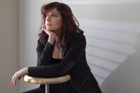 Woman with dark red curls in a black blazer seated against a white slatted wall, resting on a brass stool.Woman in a gold sequined dress seated on a geometric block, dark studio background scattered with warm points of light.