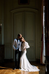 Bride and groom sharing an intimate moment in a grand European interior with tall doors and soft window light, captured in a timeless fine art wedding portrait.