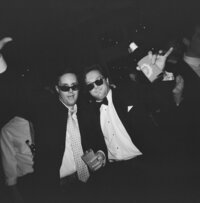 Black and white film photo of two male wedding guests wearing sunglasses, dancing and celebrating at the reception with drinks in hand.