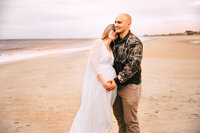 A pregnant woman laughs, leaning into her husbands neck. He's smiling brightly as they both lay their hands on her bump. They are on a beautiful golden beach.