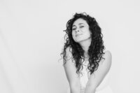 A black and white picture of a woman with dark curly hair smiling cutely against a white backdrop. She wears a long white dress and tilts her head to one side.