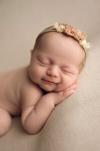 A Caucasian baby lays on a tan blanket and smiles with her eyes closed.