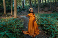 Pregnant woman in a flowing rust orange gown and crown standing on a forest path during a maternity photography session at Mill Creek Park