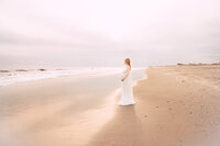A wide photograph of a beautiful bright beach with a pregnant woman staring off to the water. She holds her bump from underneath and wears a long white dress.
